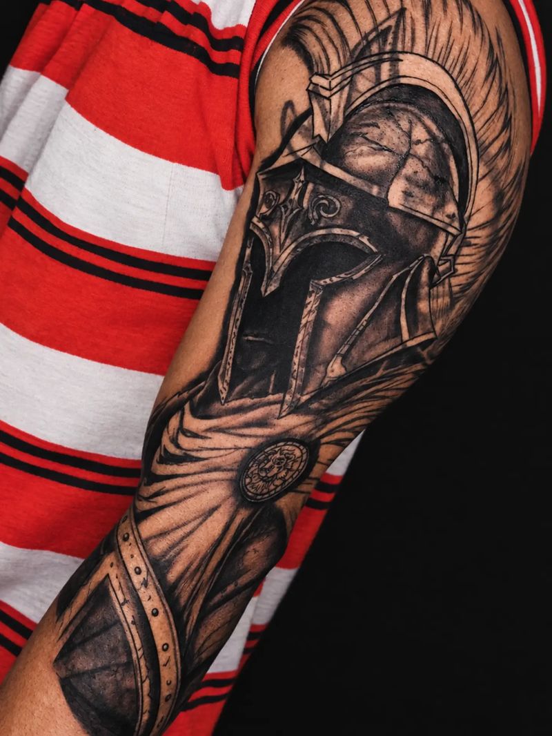Tatuagem em preto e cinza de elmo espartano com capa e escudo no braço, por Davi Pimentel Tattoo