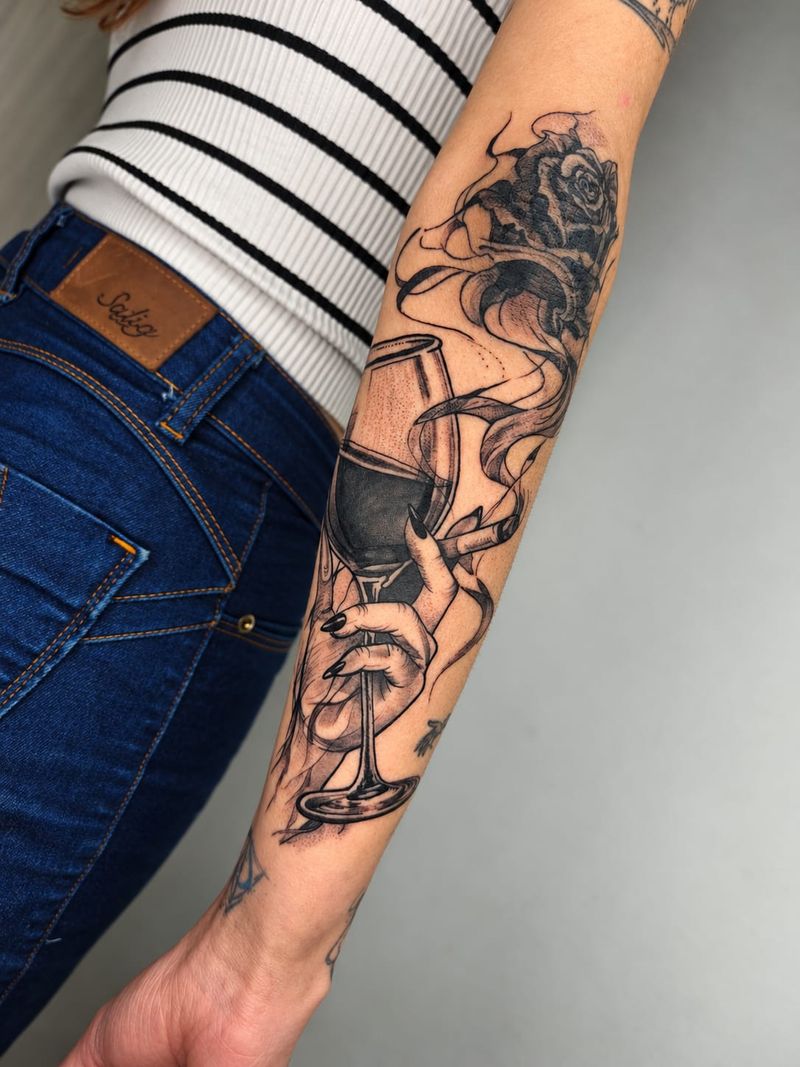 Tatuagem em preto e cinza de mão segurando taça de vinho e cigarro, com rosa e fumaça, no antebraço